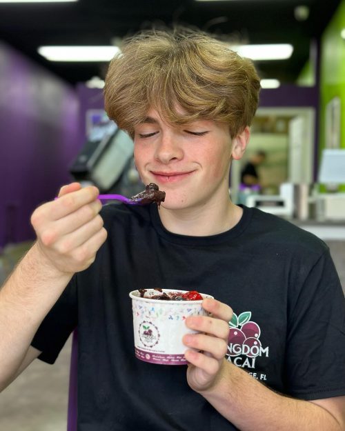 A smiling teenage boy with blond hair holds a cup of the best acai in Florida, lifting a spoonful to his mouth. He is wearing a black t-shirt with a fruit logo, sitting in a brightly colored cafe.