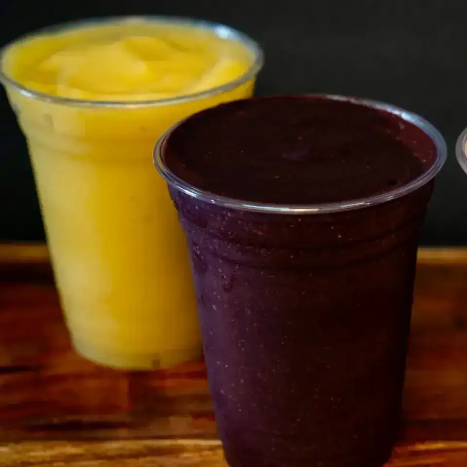Two plastic cups filled with smoothies sit on a wooden surface; one is bright yellow, and the other, featuring the best acai in Florida, is a deep purple color.