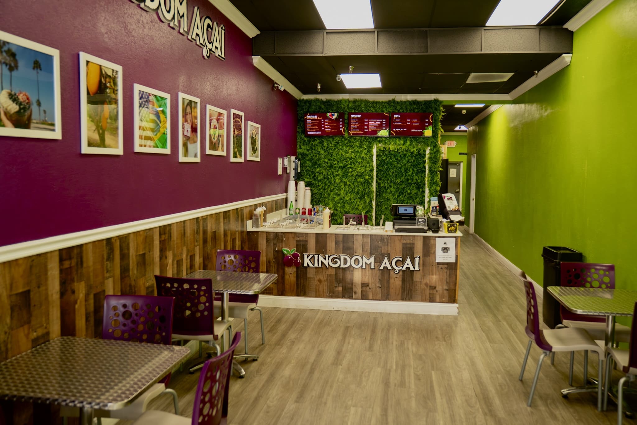 A bright, modern caf&eacute; with purple and green walls, wooden wainscoting, metal tables, and purple chairs. Enjoy the best acai in Florida at the counter with a green plant backdrop, menu board, and Kingdom A&ccedil;a&iacute; sign. Framed photos hang on the wall.