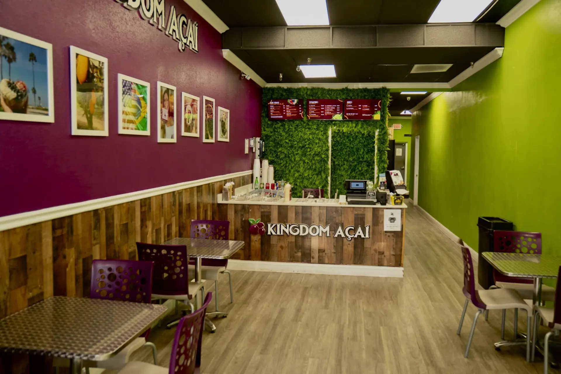 A bright, modern café with purple and green walls, wooden wainscoting, metal tables, and purple chairs. Enjoy the best acai in Florida at the counter with a green plant backdrop, menu board, and Kingdom Açaí sign. Framed photos hang on the wall.