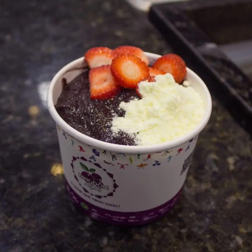 A bowl of açaí topped with sliced strawberries and white powder sits on a dark countertop. The colorful container hints at the best acai in Florida, perfect for anyone searching for the best acai near me.