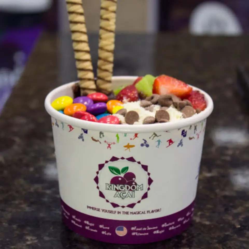 A cup of açaí from Kingdom Açaí, topped with strawberries, kiwi, colorful candy-coated chocolates, chocolate chips, shredded coconut, and two wafer sticks on a dark countertop—experience the best acai in Florida.